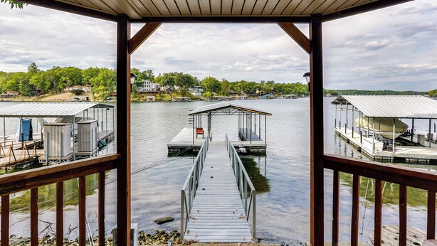 A wooden dock leading to a covered boat launch area with a view of a calm lake and houses in the background. - Lakefront Home w/ Private Dock in Sunrise Beach! (Sunrise Beach)