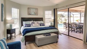 A bedroom with a bed, bedside tables, a bench, and a view of an outdoor patio with a pool and dining area.