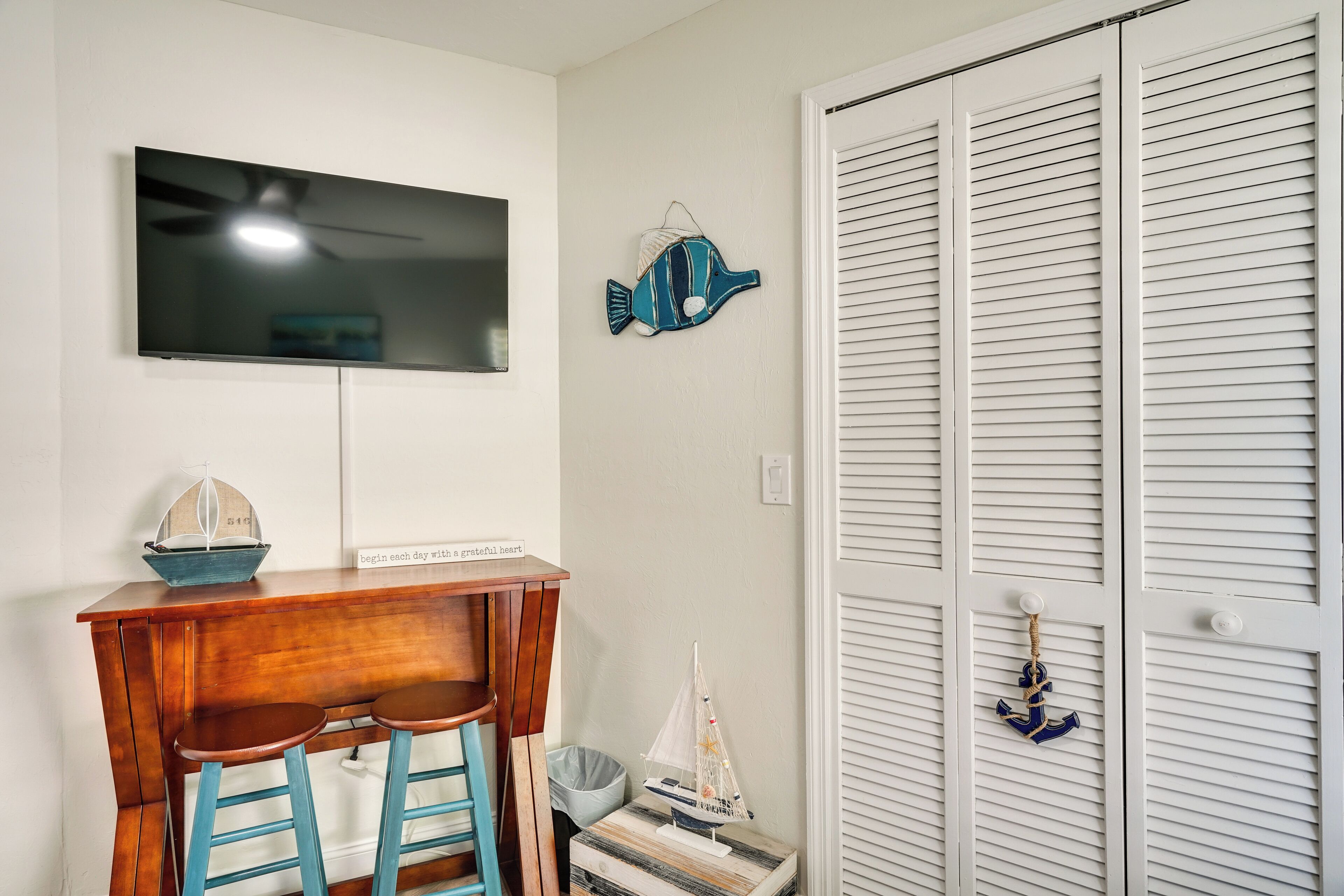 Una habitación con una televisión en la pared, una pequeña mesa de madera con dos sillas, un pez decorativo y una puerta blanca cerrada con un pequeño adorno de ancla.