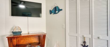 Una habitación con una televisión en la pared, una pequeña mesa de madera con dos sillas, un pez decorativo y una puerta blanca cerrada con un pequeño adorno de ancla.