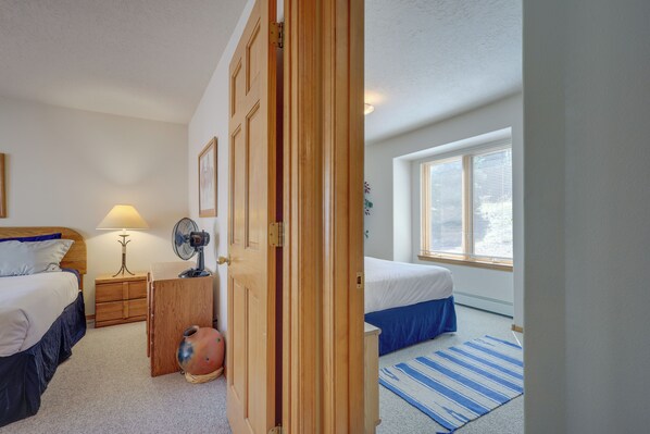 A bedroom with two beds, a wooden wardrobe, a bedside table with a lamp, a fan, and a window with blinds.