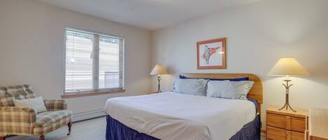 A bedroom with a large bed, a chair, a nightstand, a lamp, and a window with blinds.