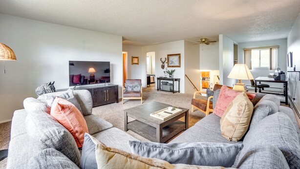 Ein modernes Wohnzimmer mit einem grauen Sofa, einem Couchtisch und einem Fernseher. Es gibt eine Essecke mit Tisch und Stühlen, und im Hintergrund befindet sich eine Küche.