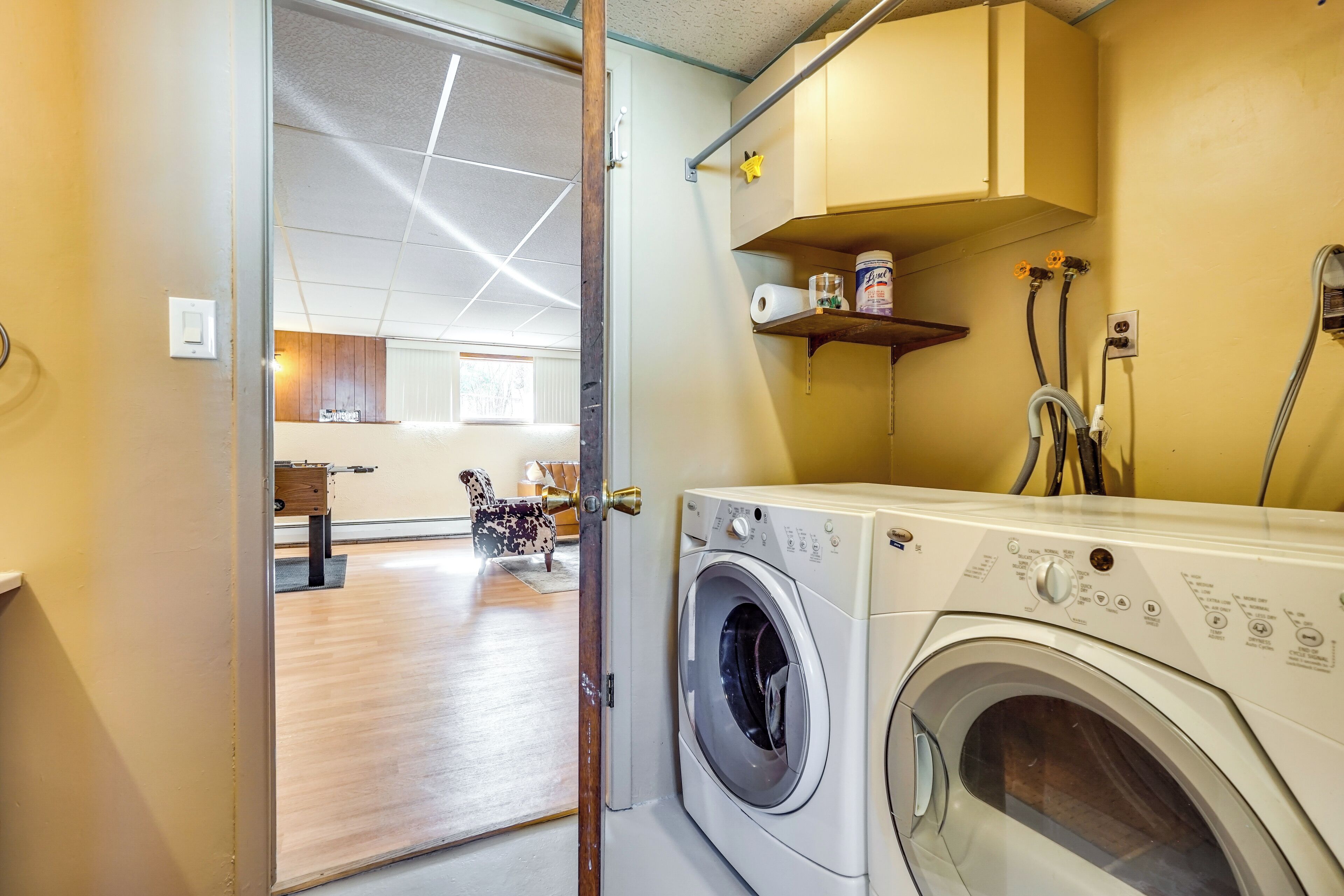 A laundry room with a front-loading washing machine, a shelf with cleaning supplies, and a door leading to a living area with a sofa and a coffee table.