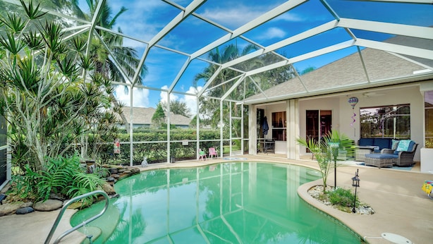 Ein Poolbereich mit Glasdach, umgeben von tropischen Pflanzen und einer Terrasse mit Gartenmöbeln.
