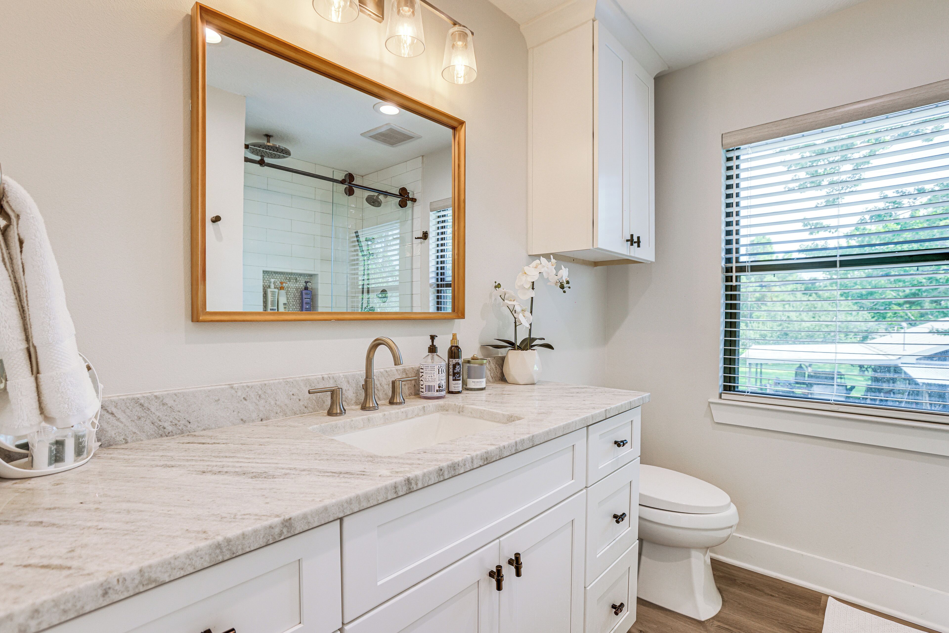 A bathroom with a marble countertop, a large mirror, a toilet, and a window with blinds.