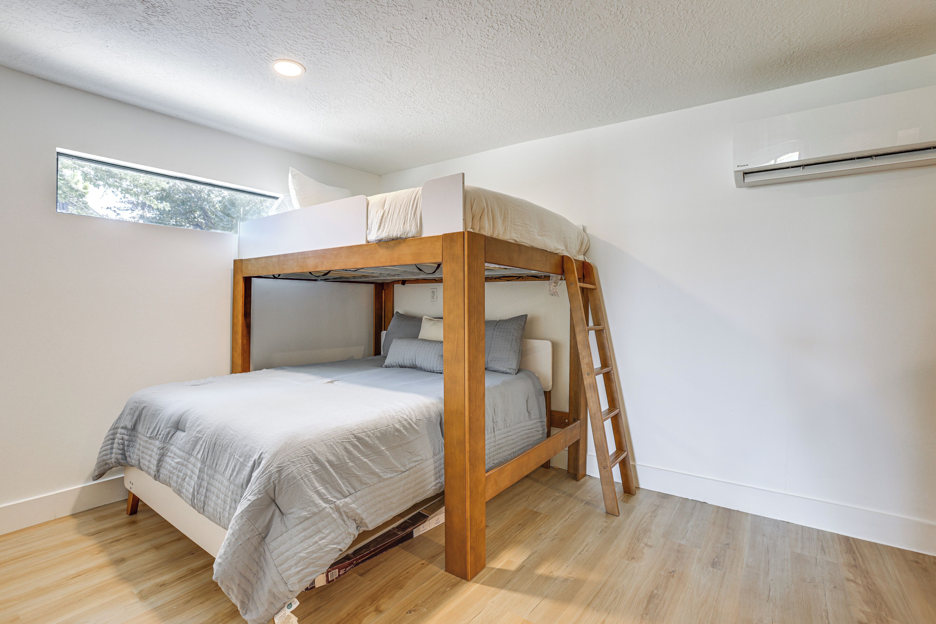 A bunk bed with a ladder, a window, and an air conditioning unit in a room with wooden flooring.