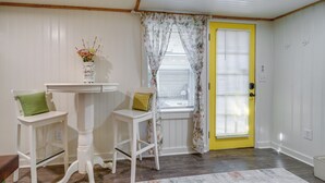 A room with a yellow door, white walls, a small table with two chairs, a vase with flowers, and a window with floral curtains. - Wilmington Area Studio ~ 4 Mi to Downtown! (Leland)