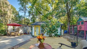 A wooden deck with a table and chairs, a small house with a yellow door, and a pink building with a tree design. - Wilmington Area Studio ~ 4 Mi to Downtown! (Leland)