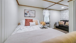 A bedroom with a bed, a nightstand, a window with curtains, a wall-mounted painting, and a sofa in the adjacent room. - Wilmington Area Studio ~ 4 Mi to Downtown! (Leland)