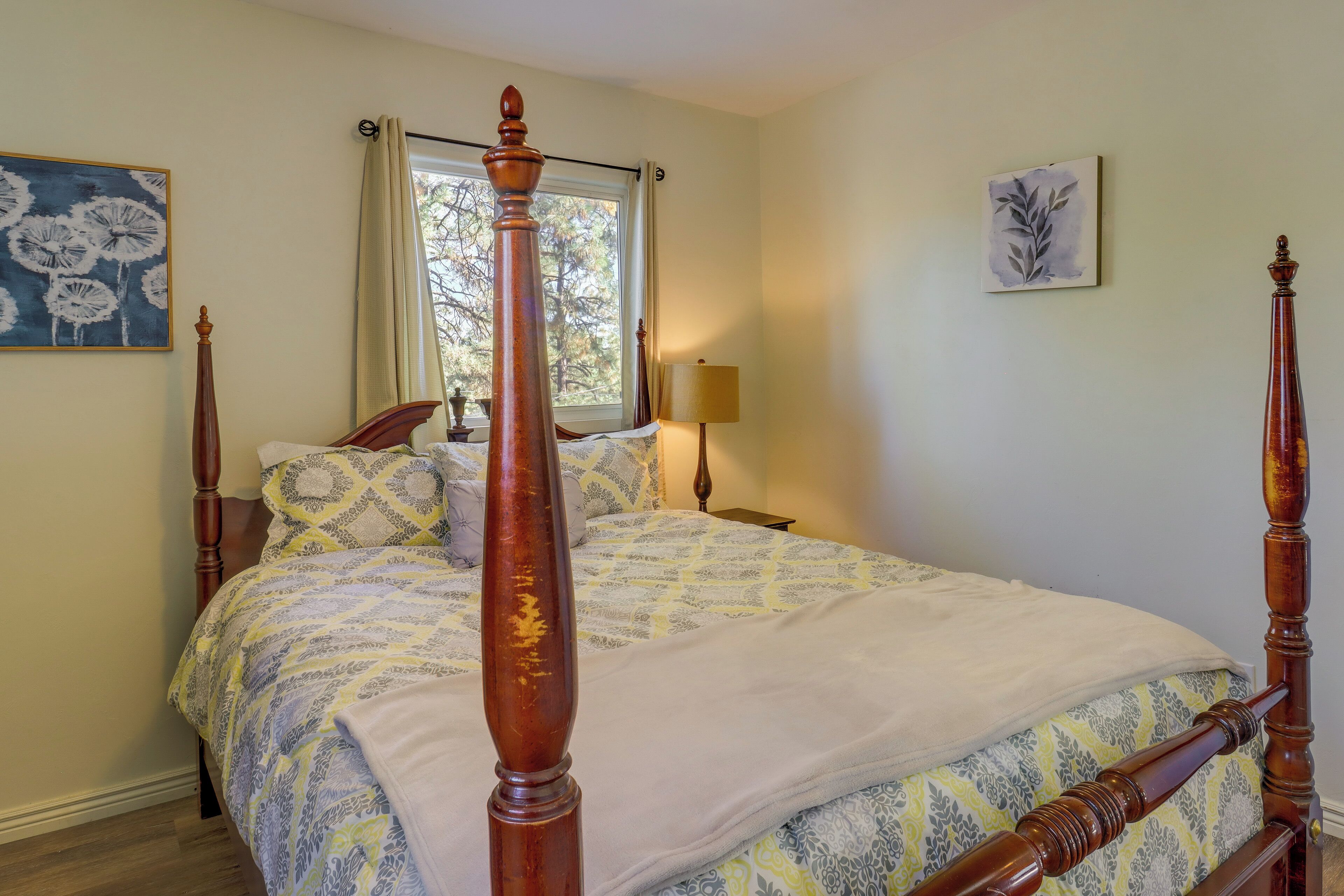 A bedroom with a four-poster bed, a window with curtains, a bedside lamp, and two wall-mounted artworks.