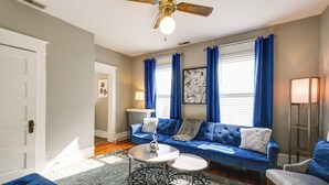 A living room with a blue sofa, two round tables, a ceiling fan, and a large window with blue curtains. - Cozy Roanoke Vacation Rental: 2 Mi to Downtown! (Roanoke)