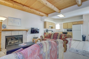 A living room with a fireplace, sofa, and television. There is a kitchen area with cabinets and appliances in the background. - Charming Amalia Retreat w/ Fireplace & Grill! (Amalia)