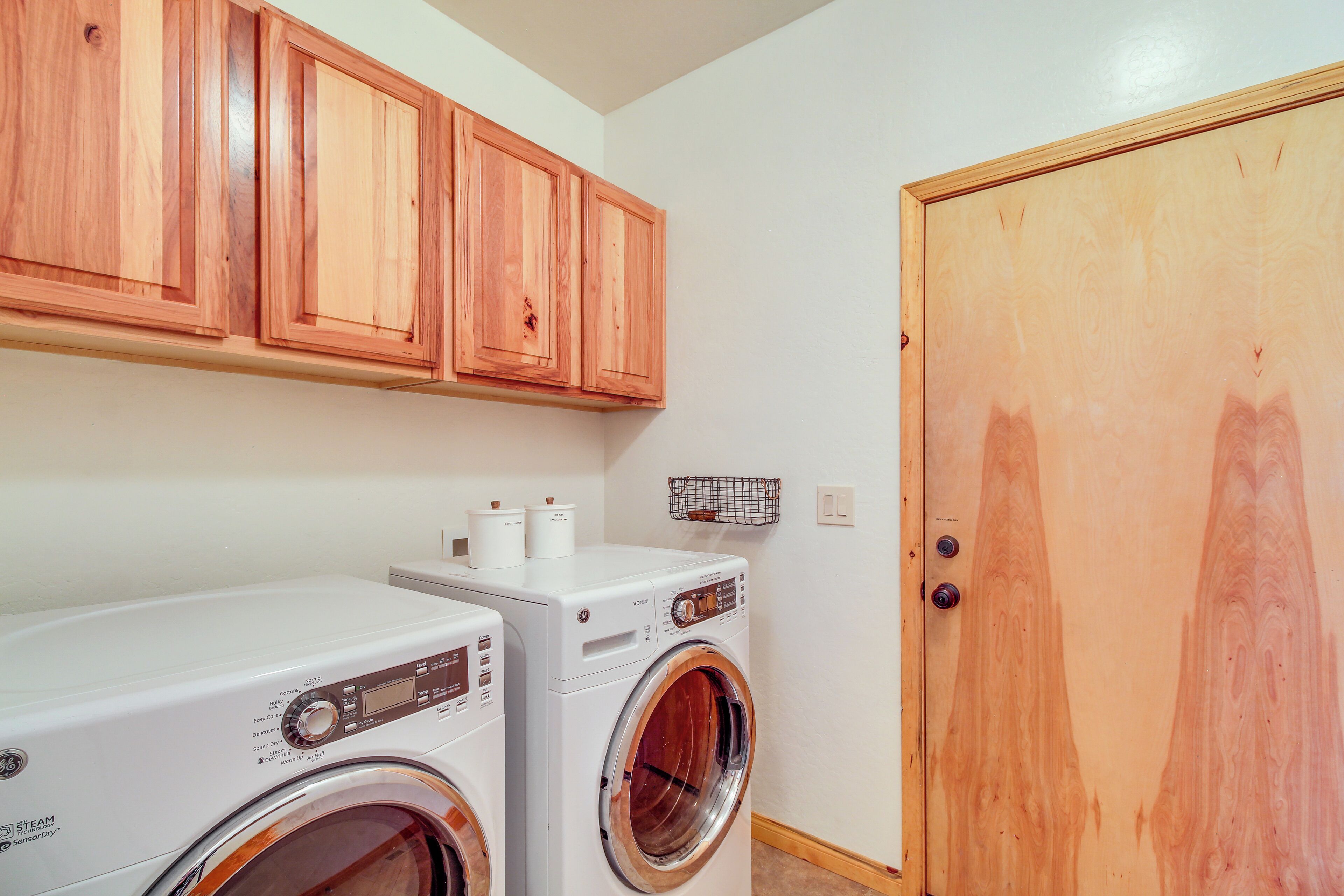A laundry room with two front-loading washing machines, wooden cabinets above, and a wooden door.