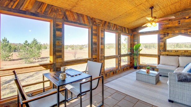 Un interior rústico con ventanas amplias, vigas de madera, un ventilador de techo y un área de estar con una mesa y sillas.