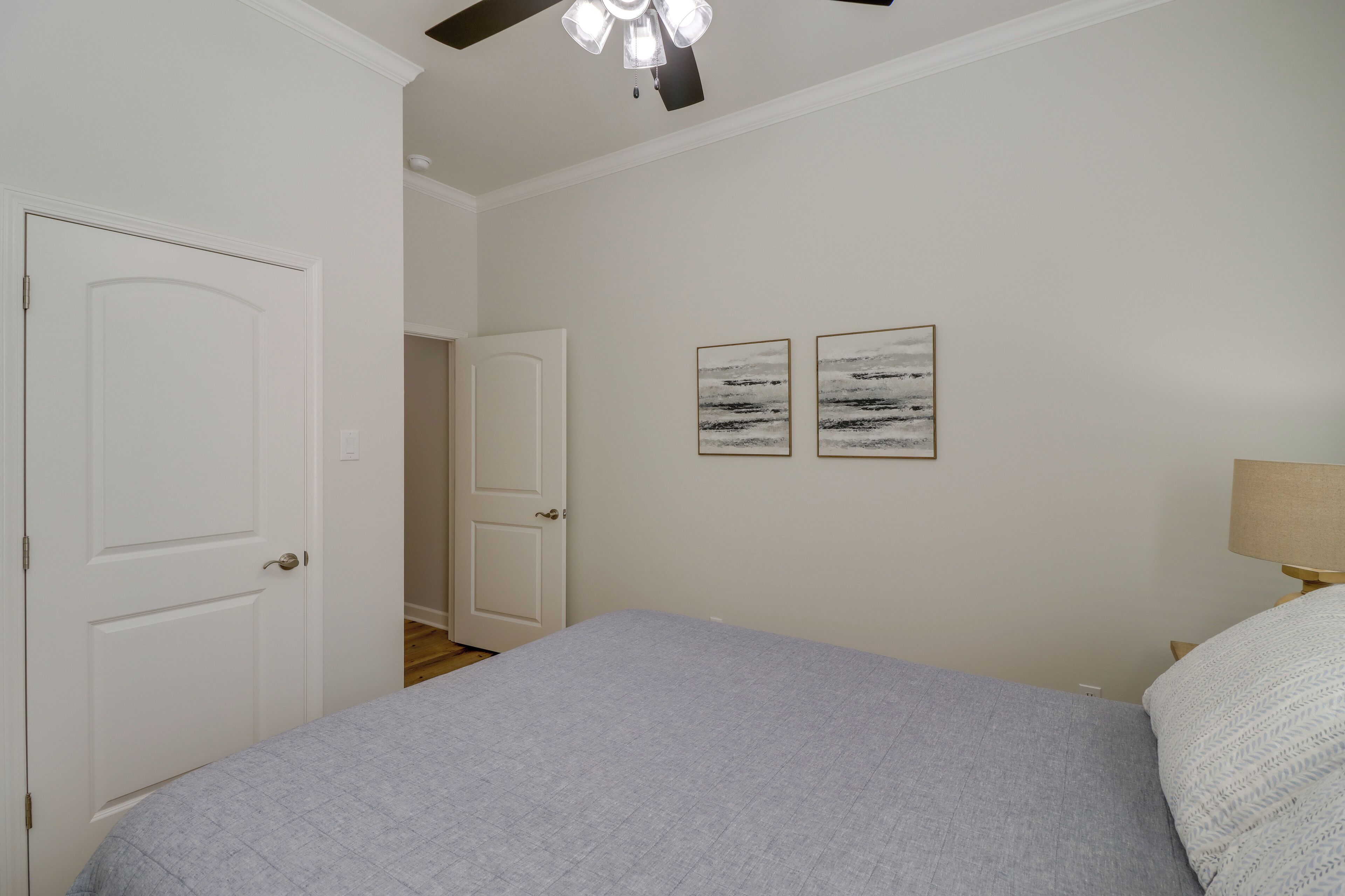 A bedroom with a bed, two framed pictures on the wall, a ceiling fan, and a door.