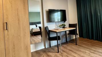 Ein modernes Hotelzimmer mit einem Holzschrank, einem Bett mit weißer Bettwäsche, einem an der Wand montierten Flachbildfernseher, einem Holztisch mit zwei Stühlen und einem grünen Vorhang.