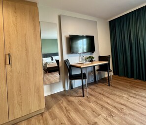 Quarto de hotel moderno com guarda-roupa de madeira, cama com roupa de cama branca, televisão de tela plana na parede, escrivaninha de madeira com duas cadeiras e cortina verde.