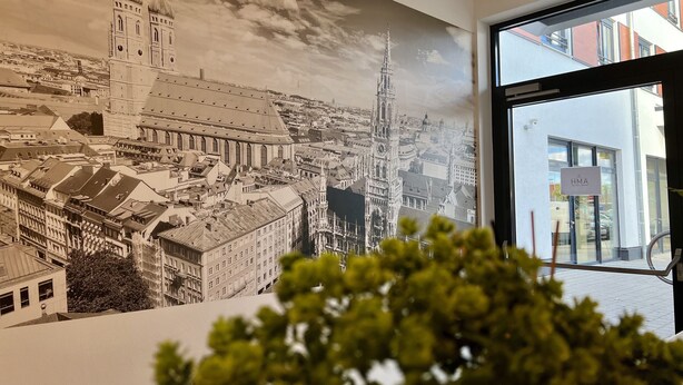 A detailed mural of a cityscape with prominent buildings and a clock tower, viewed from inside a building with a glass door and a visible exit sign. - AMH - Wohnen auf Zeit (Munich)