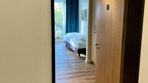 A hotel room with a wooden door, a number sign '207', a bed, a window with curtains, and a view of greenery outside. - AMH - Wohnen auf Zeit (Munich)