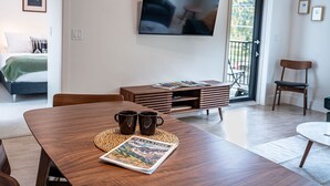 Una sala moderna con una mesa de comedor de madera, dos tazas de café y una revista. Un televisor de pantalla plana está montado en la pared, y hay un balcón con vistas a árboles.