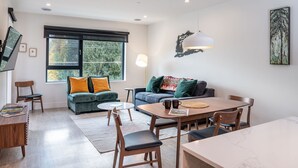 Una sala de estar moderna con sofá, mesa de centro y mesa de comedor. Hay un televisor montado en la pared, una ventana con vista a árboles y una lámpara.