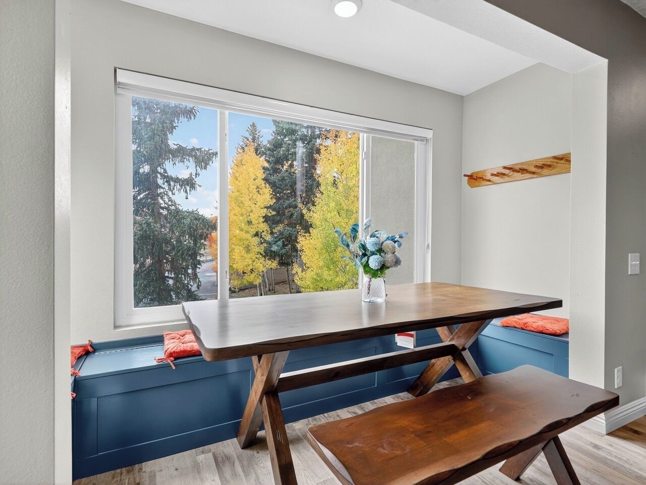 A dining area with a table and bench, a window with a view of trees, and a vase with flowers.
