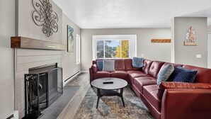 A living room with a fireplace, a sectional sofa, a coffee table, and a large window with a view. - Snowy Peaks Haven | Walk to Slopes & Park (Brian Head)