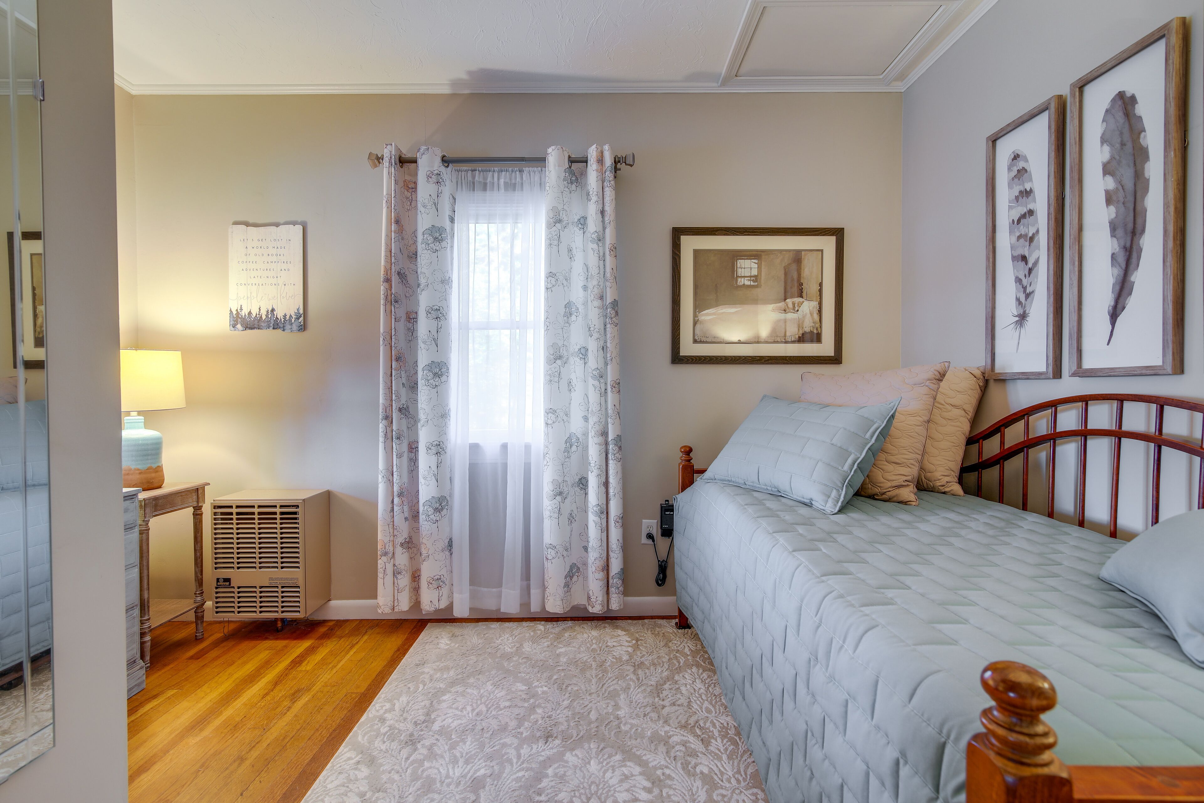 Een slaapkamer met een bed, nachtkastje, lamp, radiator en een raam met bloemen gordijnen.