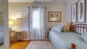 Een slaapkamer met een bed, nachtkastje, lamp, radiator en een raam met bloemen gordijnen.