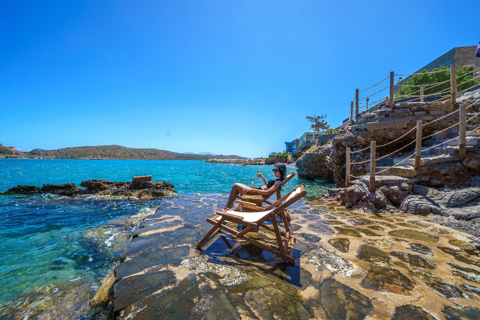 A wooden chair on a rocky shore with clear blue water and a stone building in the background.