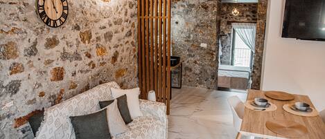 Una habitación acogedora con pared de piedra, separador de madera, sofá con cojines estampados y una mesa de comedor de madera con un televisor en la pared.
