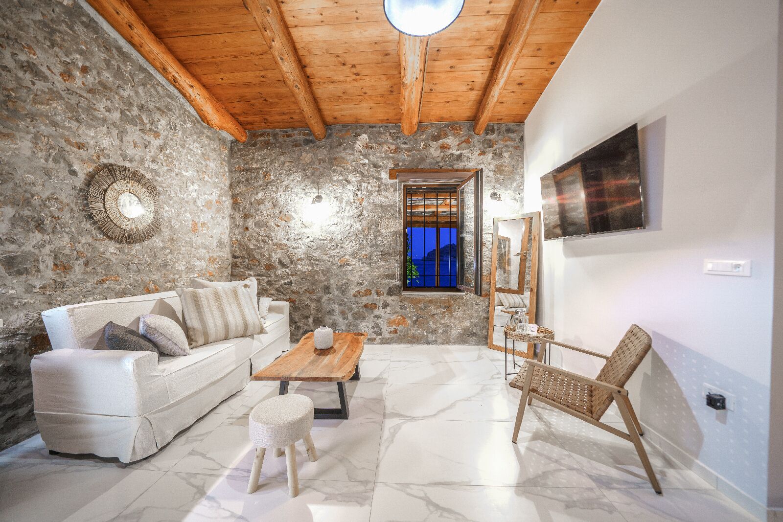 Un salon chaleureux avec un mur en pierre, des poutres apparentes en bois au plafond, un canapé blanc, une table basse en bois, un fauteuil en osier et une vue sur la mer par la fenêtre.