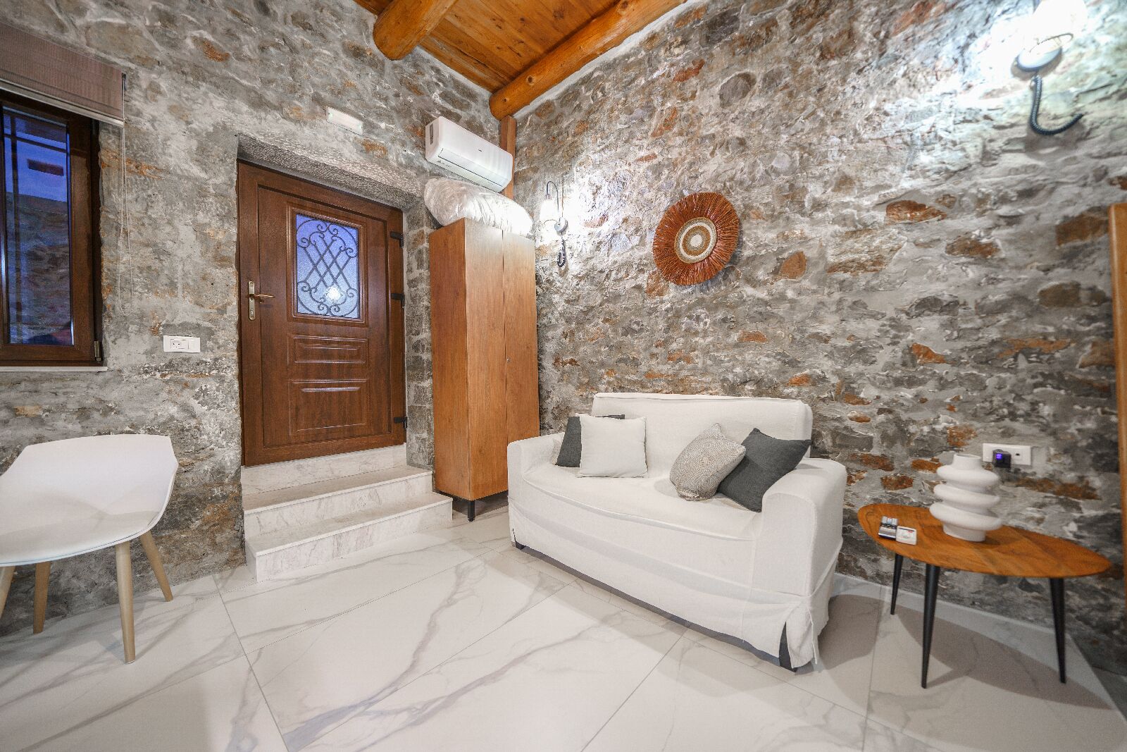 A room with a stone wall, a wooden door, a white sofa, a white chair, and a small table with a lamp.