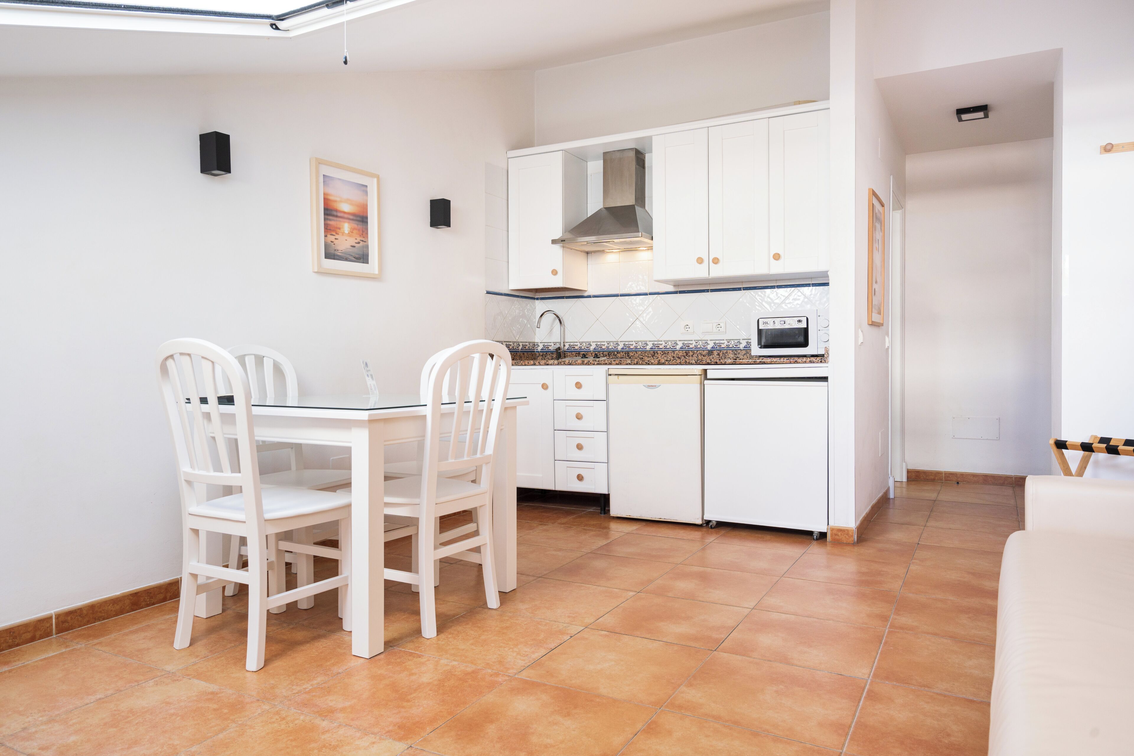 Cocina compacta con armarios blancos, una mesa de comedor con sillas y piso de baldosas.