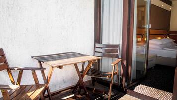 Un balcón con una mesa y sillas de madera, y una puerta corrediza de vidrio que da a un dormitorio con cama y mesita de noche.