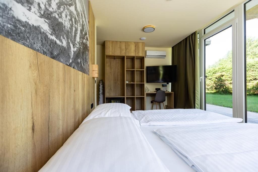 Een hotelkamer met een bed, een bureau, een televisie en een groot raam met uitzicht op groen.