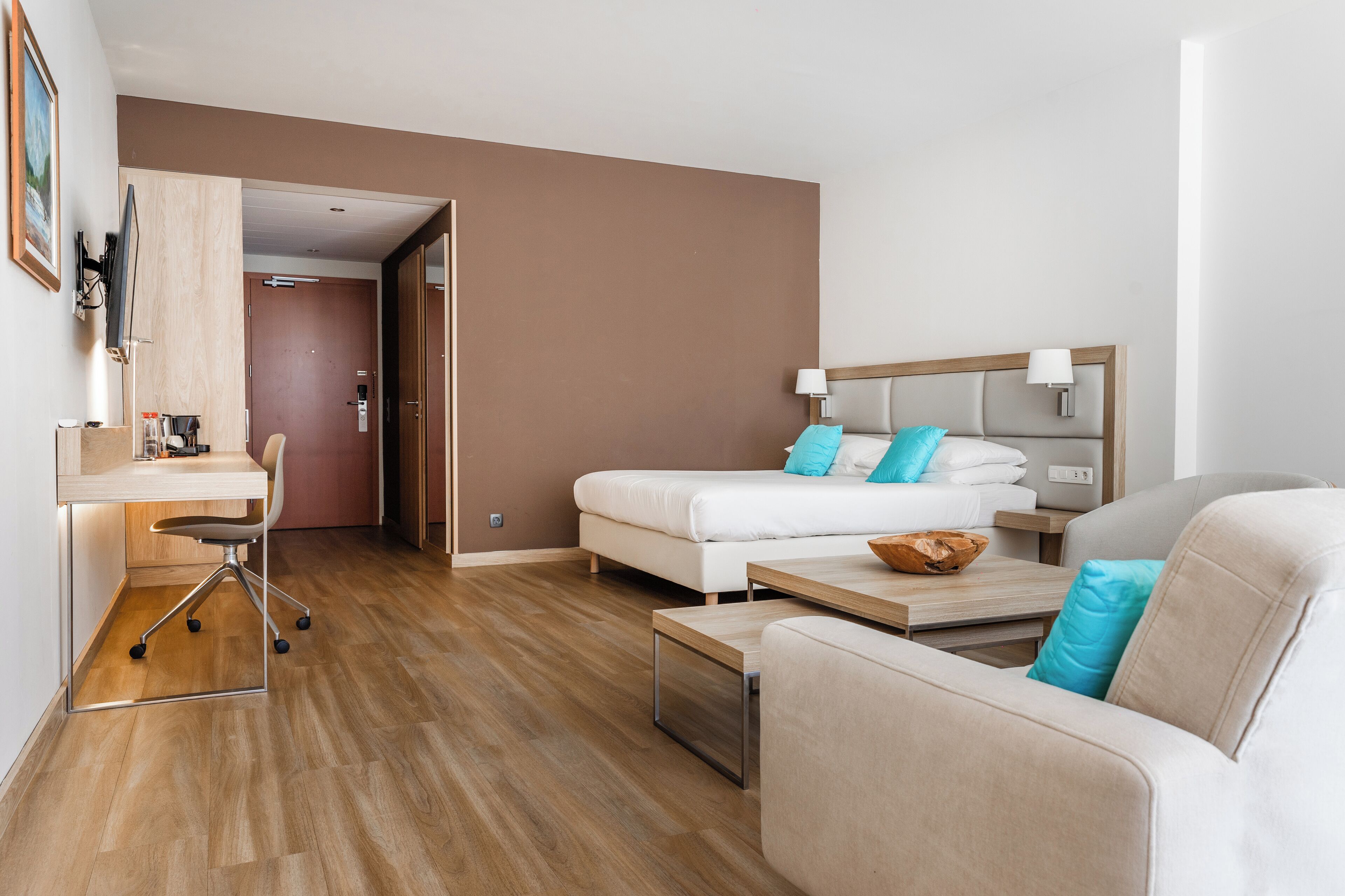 A modern hotel room with a wooden floor, a white bed with blue pillows, a beige sofa, a desk with a chair, and a small table with a bowl.