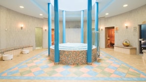 A large indoor pool area with a central hot tub, surrounded by blue pillars, and a tiled floor with a geometric pattern.