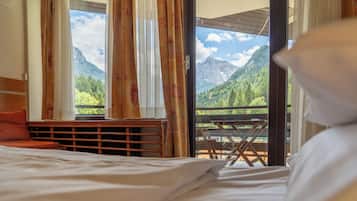 A hotel room with a bed, a view of mountains through the window, and a balcony with a table and chairs.