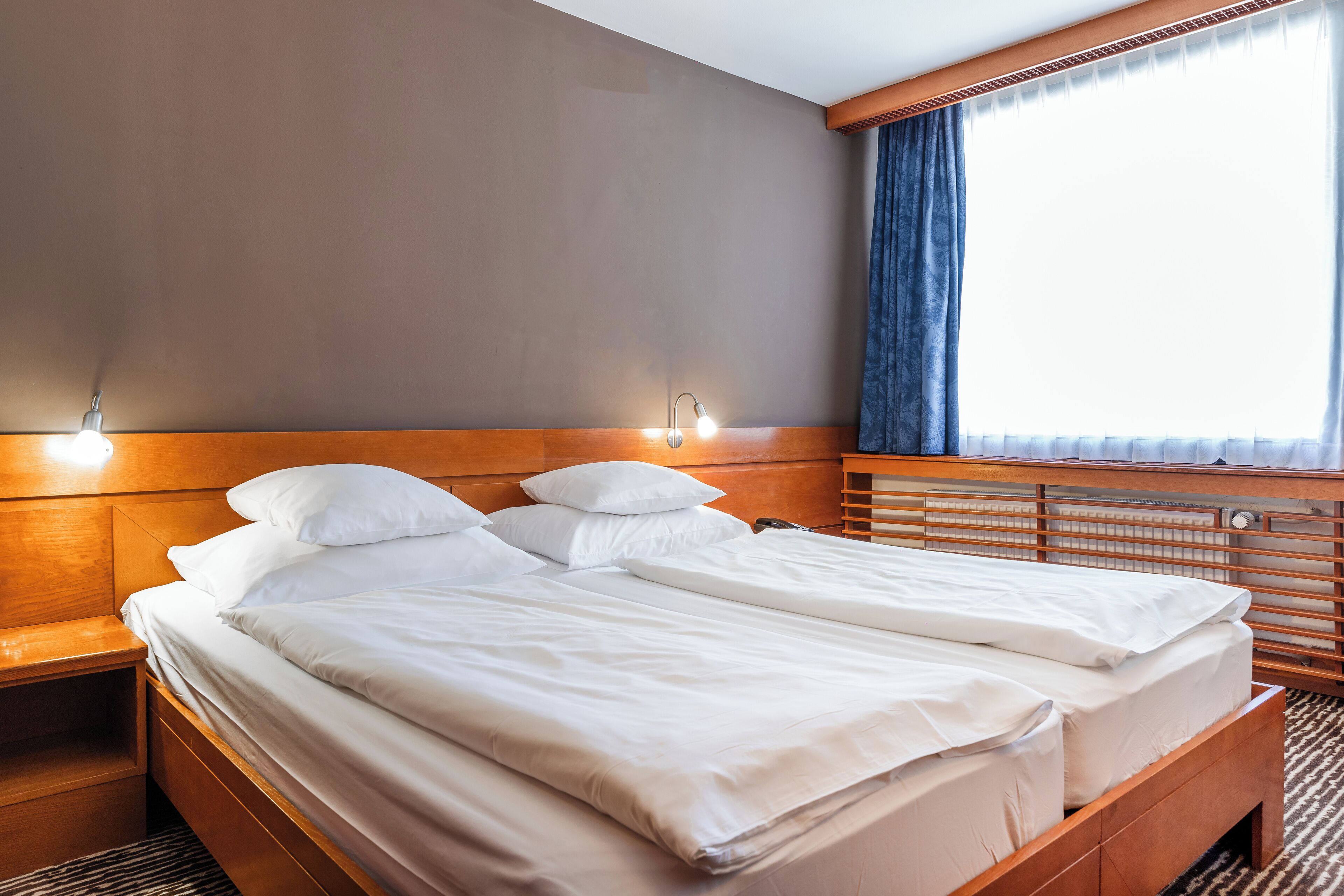A hotel room with a double bed, wooden headboard, bedside tables, and a large window with curtains.