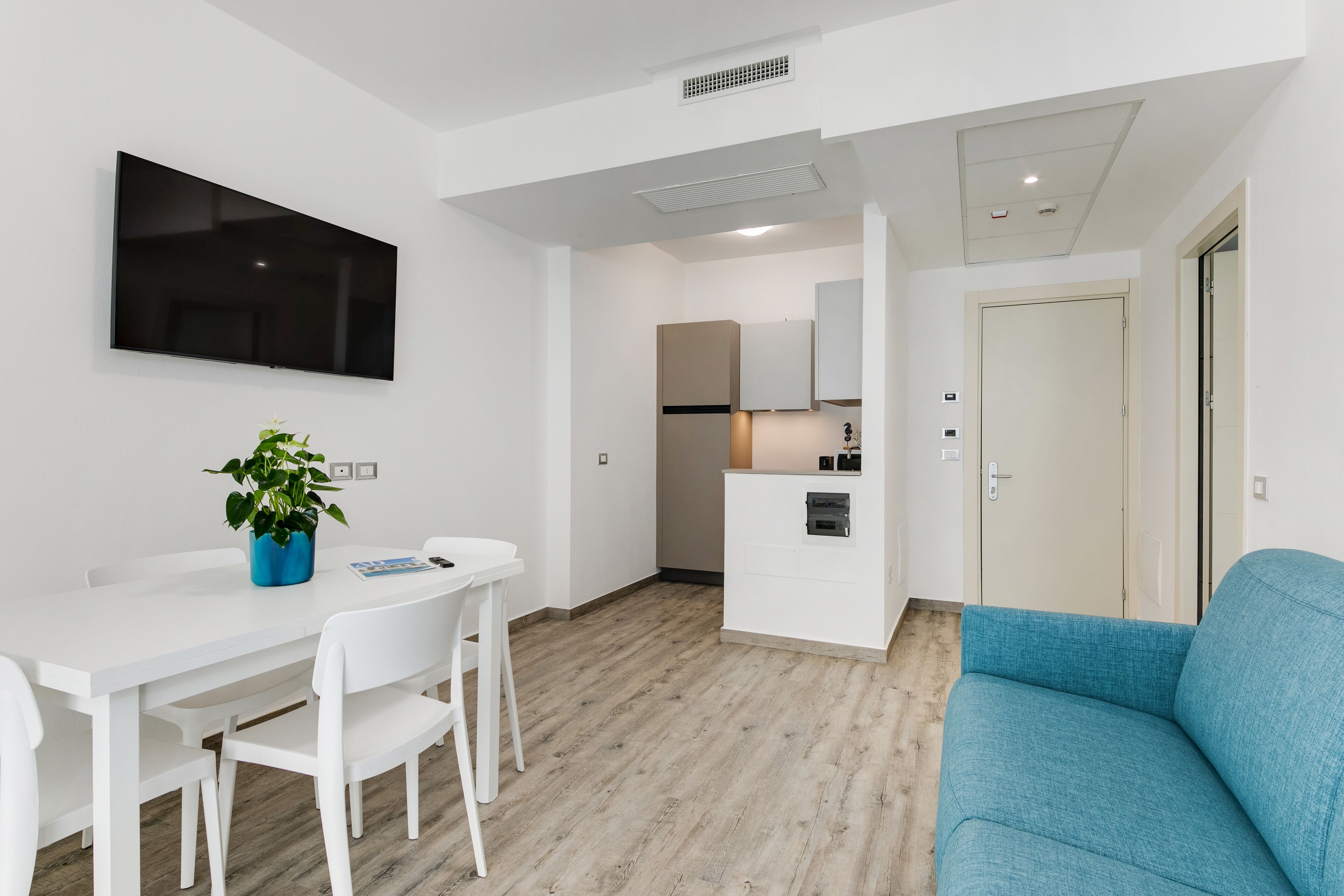 Een moderne, goed verlichte kamer met een witte eettafel, stoelen, een blauwe bank en een keuken op de achtergrond.