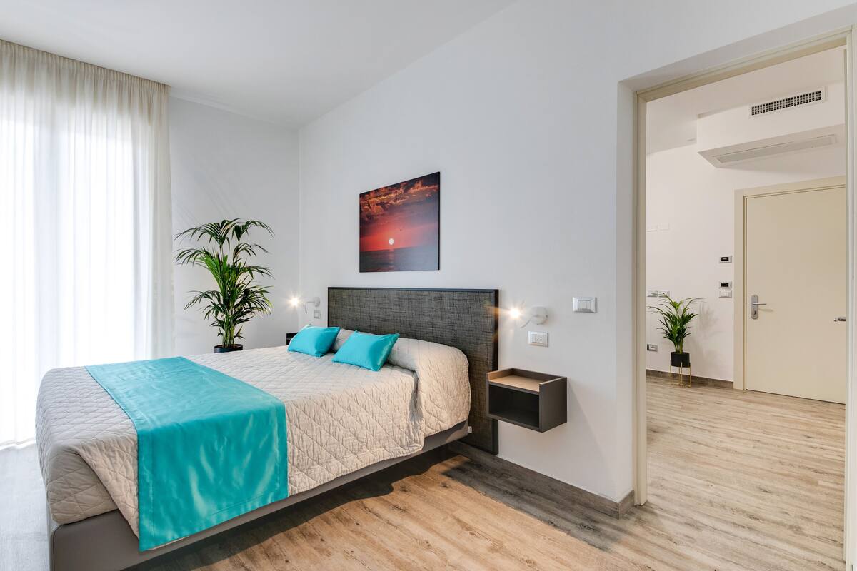 A modern bedroom with a large bed, a nightstand, a wall-mounted shelf, and a potted plant.