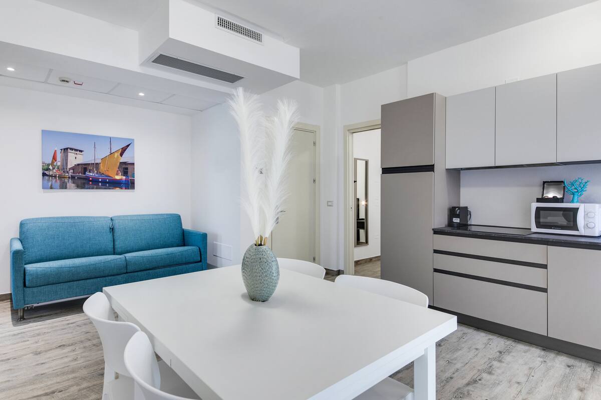 A modern living room with a blue sofa, a white dining table, and a kitchen area with a microwave.