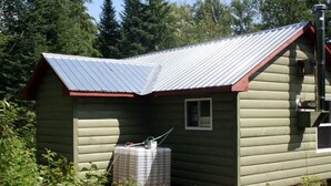 A log cabin with a metal roof, a small window, and a water tank in the foreground. - Hunting camp #10 (Egan-Sud)