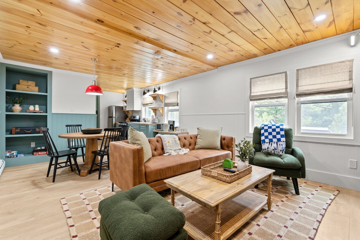 Una sala de estar acogedora con un sofá marrón, un sillón verde y una mesa de centro de madera. Hay una zona de comedor con una mesa y sillas, y una cocina al fondo.