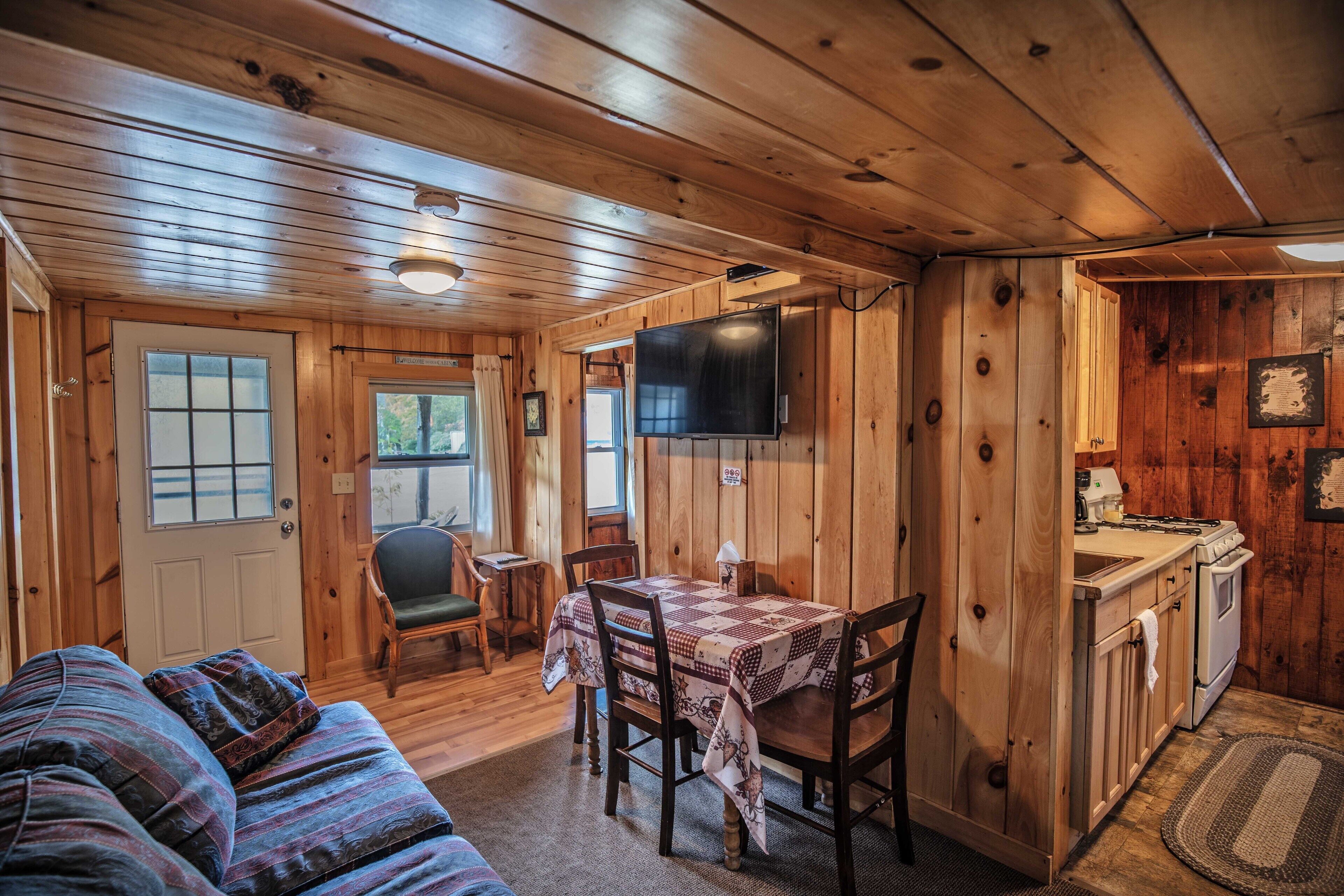 A cozy cabin interior with a wooden floor, a dining table with chairs, a sofa, a television, and a kitchenette.