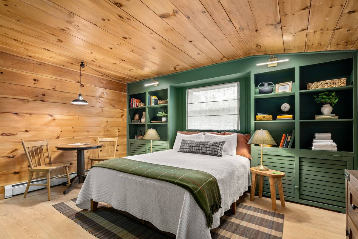 Un dormitorio acogedor con techo de madera, una cama con manta verde, una mesita con lámpara y una estantería con diversos objetos.