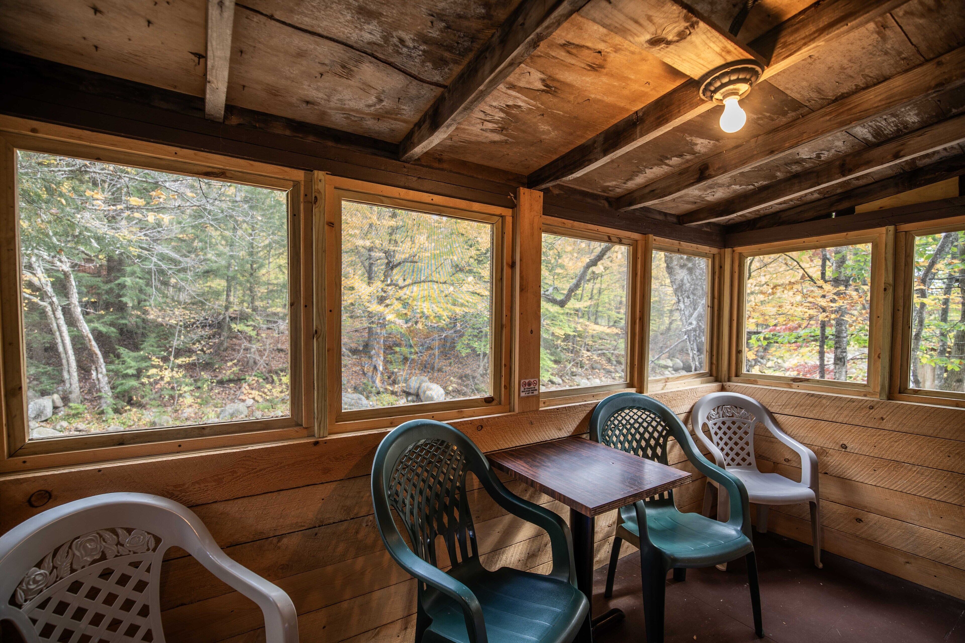 A wooden cabin interior with large windows, a table, and two chairs.