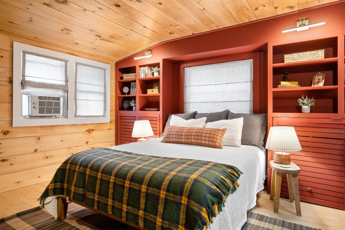 Un dormitorio acogedor con cama, lámparas de noche, una ventana con persianas y estantes empotrados con diversos objetos.